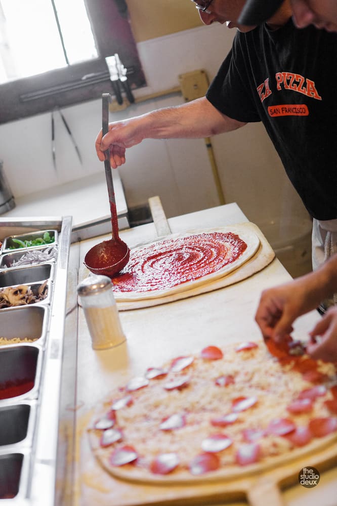 Ladling tomato sauce onto a pizza, with a pepperoni pie in front